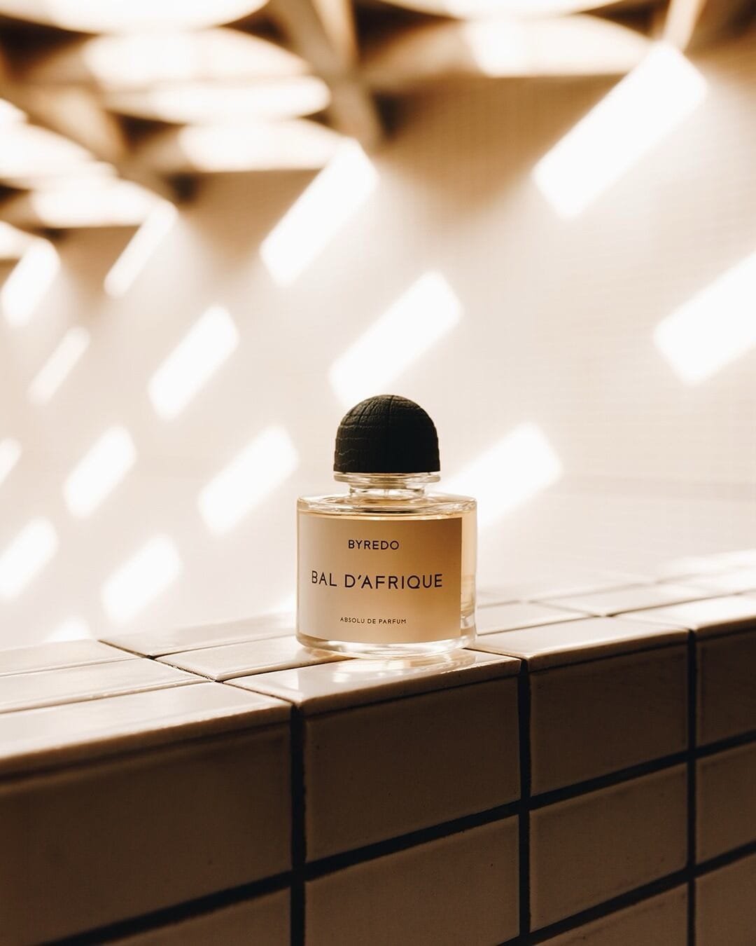 Byredo fragrance product photo: clear glass, matte black cap and pale gold, glass and stone, natural light
