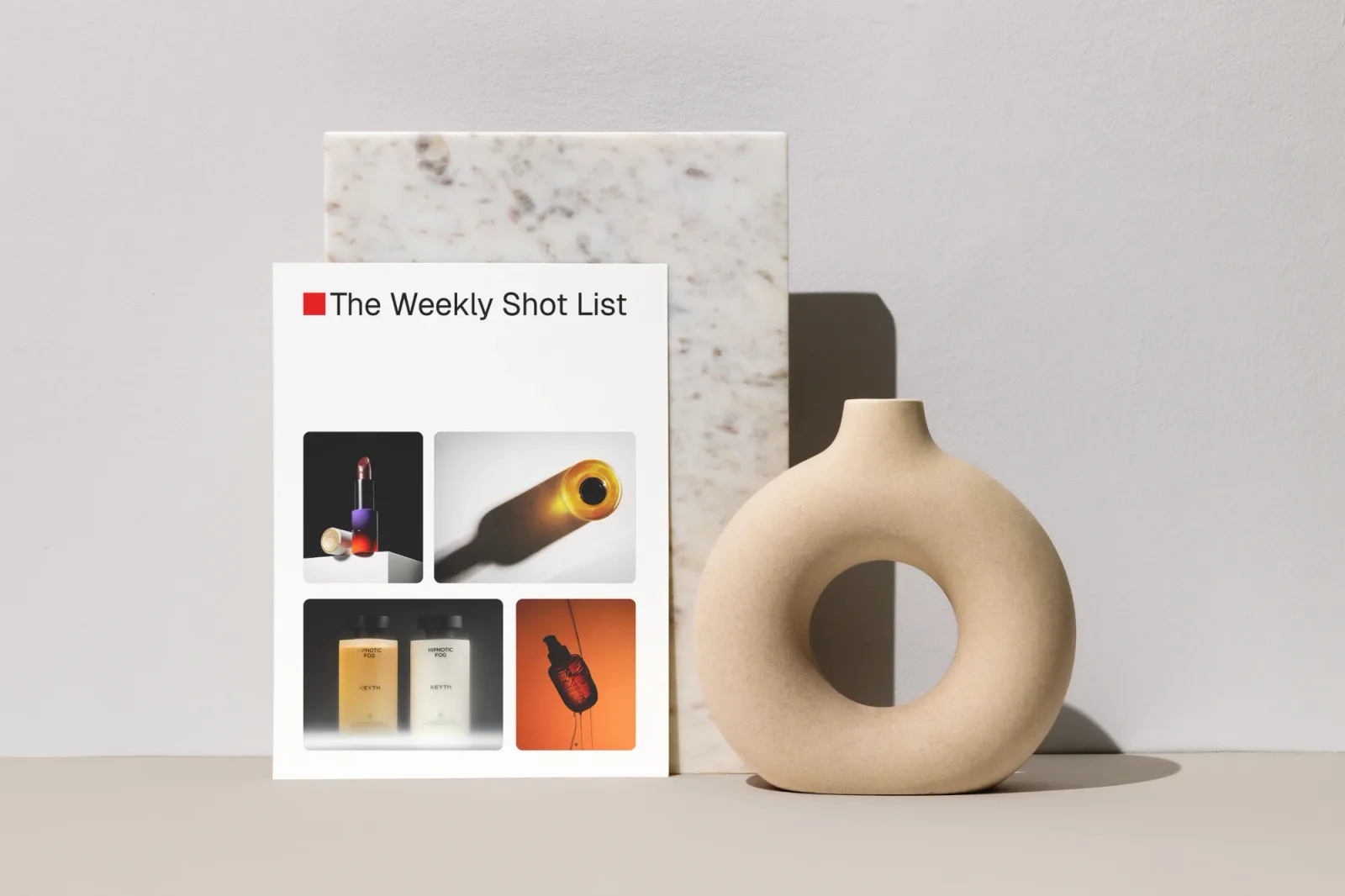 The Weekly Shot List printed on a card, leaning on marble beside a ceramic vase